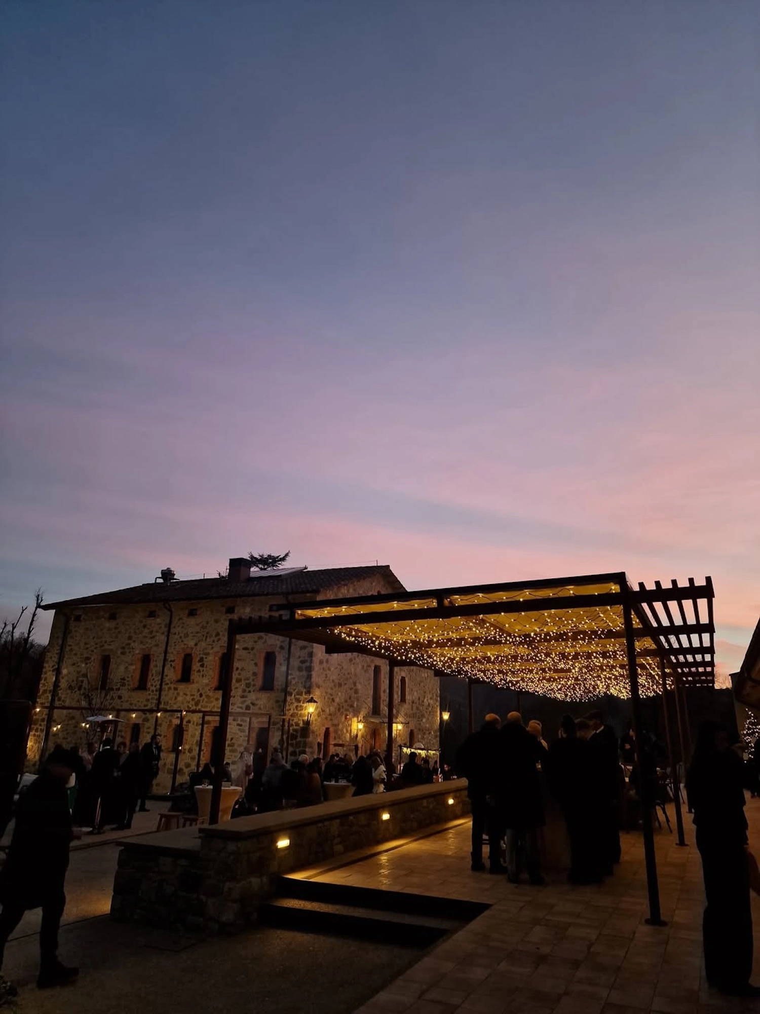 Convidats gaudint d'una celebració al capvespre sota una pèrgola il·luminada a l'exterior de la masia rural Mas Pairó.
